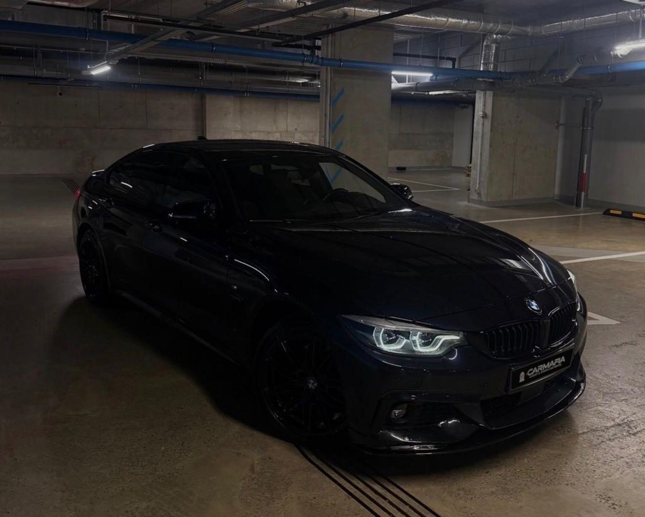 Czarne BMW nocą w podziemnym garażu, widok z przodu na reflektory i tablicę z logo CarMafia.fun